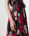 Close-up of a person wearing a dark floral skirt with large red, pink, cream, and light blue flowers, paired with a dark brown satin top against a light gray studio background.