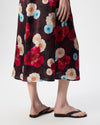 A person wearing a dark floral midi skirt with large red, cream, and light blue flowers, paired with black flip-flop sandals, shown from the waist down against a plain white studio background.