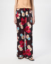A model wearing wide-leg trousers with a dark burgundy base and a vibrant multicolor floral print in red, pink, blue, and cream, paired with a beige top and black sandals against a white studio background.