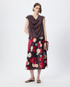 A model stands against a light gray studio background wearing a dark brown draped sleeveless top and a black midi skirt with a vibrant red, pink, and white floral pattern.