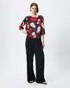 Model wearing a dark burgundy patterned smock top with floral print and black trousers against a white/grey background. 