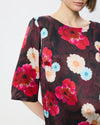 Model wearing a dark burgundy patterned smock top with floral print and black trousers against a white/grey background. 