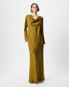 A woman modeling a long-sleeved, floor-length draped dress in a rich mustard yellow color, set against a plain light gray studio background.