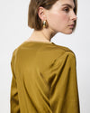 A woman shown from the back and side wearing a satin-like olive-gold top and chunky gold hoop earrings against a plain light gray background.