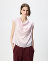 A woman modeling a light pink sleeveless cowl-neck top with a soft satin-like sheen, paired with dark burgundy trousers, against a plain white studio background.