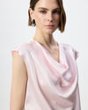 A close-up of a model wearing a light pink satin top with a draped cowl neckline and cap sleeves, set against a plain light gray background.