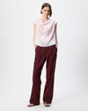 A model stands against a light gray studio background wearing a sleeveless draped light pink top and dark burgundy wide-leg trousers with black sandals.