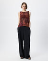 A model stands against a light gray studio background wearing a sleeveless top with an abstract rust, burgundy, and brown pattern on the upper half and black wide-leg pants.