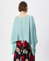 A model shown from the back wearing a light blue-green draped poncho-style top over a dark floral skirt, against a plain white studio background.