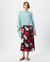 A model stands against a light gray studio background wearing a light blue draped top and a dark midi skirt with a red, pink, and blue floral pattern.