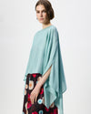 A woman wearing a flowing light blue draped top and a dark floral skirt, photographed against a plain light gray studio background.
