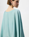A woman shown from the back and side wearing a flowing light aqua blue cape-style top against a plain light gray studio background.
