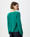 A woman with short brown hair is shown from the back wearing a teal green long-sleeve blouse and dark navy trousers, photographed against a clean white studio background.