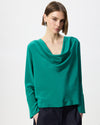 A woman wearing a teal green long-sleeve cowl neck blouse with dark trousers, photographed against a clean white studio background.