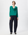 A model stands against a light gray studio background wearing a bright green long-sleeve cowl-neck top and dark navy wide-leg trousers.