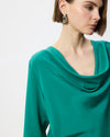 A close-up of a woman wearing a vibrant emerald green cowl-neck top and matching green teardrop earrings against a plain white background.