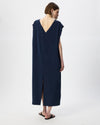 A woman shown from the back wearing a sleeveless navy maxi dress with a deep V-neckline and a front slit, paired with black slide sandals against a plain white studio background.