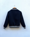 A navy varsity-style jacket with white striped ribbing on the collar, cuffs, and hem, hanging on a wooden hanger against a white textured wall.