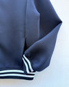 Close-up of a navy blue textured jacket sleeve with white and navy striped ribbed cuffs, shown against a plain off-white background.