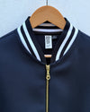 A close-up of a dark navy blue textured zip-up jacket hanging on a wooden hanger against a white textured wall. The jacket features a ribbed collar with white and light blue stripes, a gold zipper.