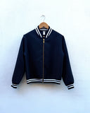 A navy blue bomber jacket with white and black striped ribbed trim, hanging on a wooden hanger against a white textured wall.