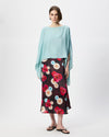 A model stands against a light gray studio background wearing a light blue draped cape-style top and a dark midi skirt with a colorful floral print in red, pink, white, and light blue, paired with black flip-flop sandals.