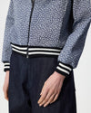 A close-up of a person wearing a cropped zip-front jacket with a blue and floral pattern, black-and-white striped ribbed hem and cuffs, and dark navy pants against a light gray studio background.
