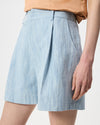 A close-up of a model wearing light blue textured shorts with a pleated front and side pockets, paired with a peach-colored top against a plain white background.