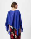 A woman shown from the back wearing a bright royal blue draped poncho-style top over dark floral pants, photographed against a plain light gray studio background.