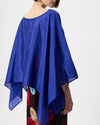 A model shown from the back wearing a vibrant royal blue satin poncho top with wide draped sleeves and an asymmetrical hem, paired with a floral skirt against a plain white background.