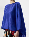 A close-up of a model wearing a vibrant royal blue satin cape-style top with wide draped sleeves, paired with a floral skirt against a light gray background.