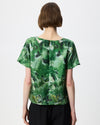 A woman viewed from the back wearing a short-sleeve green abstract print top and black pants against a plain light gray studio background.