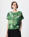 A woman modeling a short-sleeve top with an abstract green and white watercolor-style print, paired with black trousers, against a plain light gray studio background.