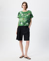 A model stands against a light gray studio background wearing a short-sleeve green abstract-print top and black shorts, with black flip-flop sandals.