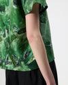 Close-up of a person wearing a green abstract patterned short-sleeve T-shirt and black shorts against a plain light background.