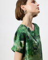 A woman wearing a green short-sleeve top with an abstract painterly print in shades of green, black, and white, shown in profile against a light gray background.