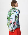 A woman shown from the back wearing a long-sleeve floral print blouse with a high collar and a bright red skirt, photographed against a plain light gray studio background.