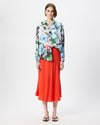 A model stands against a plain white studio background wearing a long-sleeve button-up shirt with a colorful floral print and a bright orange-red midi skirt.