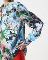 A close-up of a person wearing a long-sleeve button-up shirt with a vibrant, multicolored floral print featuring large light blue, white, and pink flowers with green leaves, paired with a red bottom.