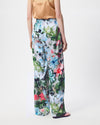 A model is wearing a pair of wide-leg trousers featuring a vibrant, multicolored floral print with white, pink, blue, and red flowers on a green and dark background. The pants have an elastic waistband and are styled with a peach satin top and black sandals against a plain white studio background.