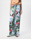 A model is wearing a pair of wide-leg trousers featuring a vibrant, multicolored floral print with white, pink, blue, and red flowers on a green and dark background. The pants have an elastic waistband and are styled with a peach satin top and black sandals against a plain white studio background.