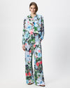 A model stands against a light gray studio background wearing a matching long-sleeve button-front top and wide-leg pants set in a light blue base fabric with a dense multicolor floral print featuring green, pink, red, and white flowers.