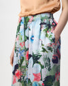 A model is wearing a pair of wide-leg trousers featuring a vibrant, multicolored floral print with white, pink, blue, and red flowers on a green and dark background. The pants have an elastic waistband and are styled with a peach satin top and black sandals against a plain white studio background.