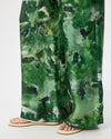 Green abstract watercolor print wide-leg pants shown on a model from the knees down, paired with beige flat sandals against a white studio background.