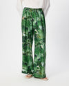 A person wearing green wide-leg pants with an abstract watercolor-style print in various shades of green, paired with a white top and beige sandals, shown against a plain white studio background.
