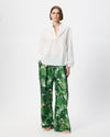 A model stands against a plain white studio background wearing a white long-sleeve blouse and green wide-leg trousers with an abstract botanical print.