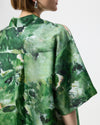 A close-up back view of a model wearing a green abstract print shirt with wide short sleeves and a high collar, set against a plain light gray background.