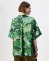 A model shown from the back wearing a green short-sleeve button-up shirt with an abstract watercolor-style print in various shades of green, paired with brown pants, against a plain white studio background.
