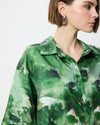 A close-up of a model wearing a green abstract print button-down shirt and silver geometric earrings against a plain white background.