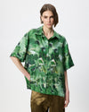 A model wearing a green short-sleeve button-up shirt with an abstract watercolor-style print in various shades of green, paired with olive green pants, against a plain light gray background.